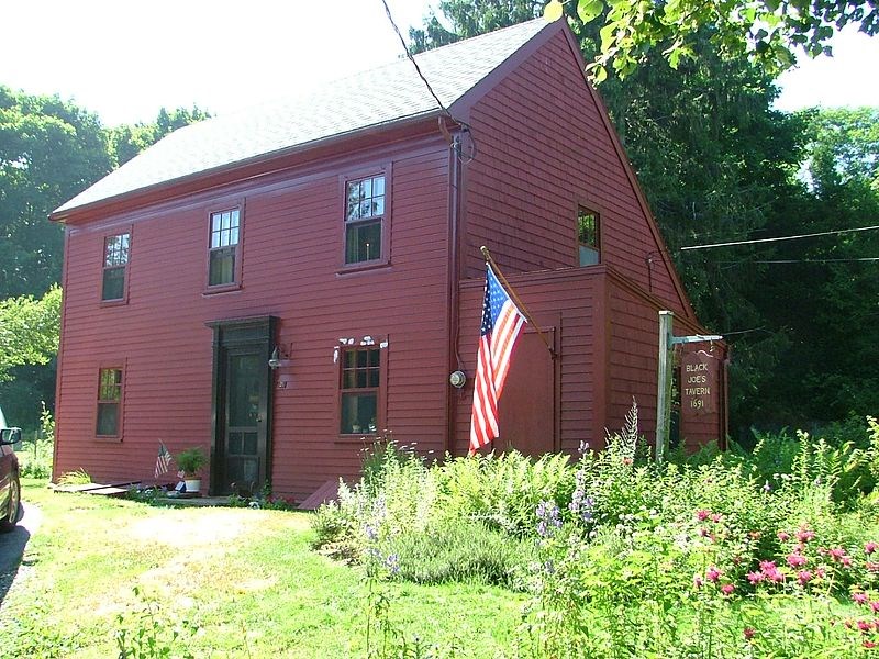 Front diagonal perspective of deep red colonial Saltbox house with black door. American flag, green grass, and wildflower garden in front. A sign reads "Black Joe's Tavern, 1691"