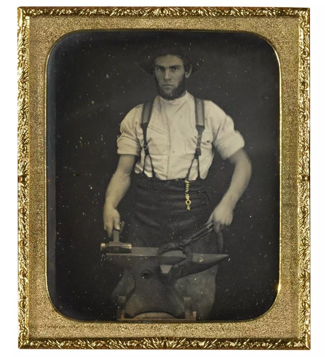 A black and white photograph of a blacksmith foraging a horseshoe within a gold metal frame.