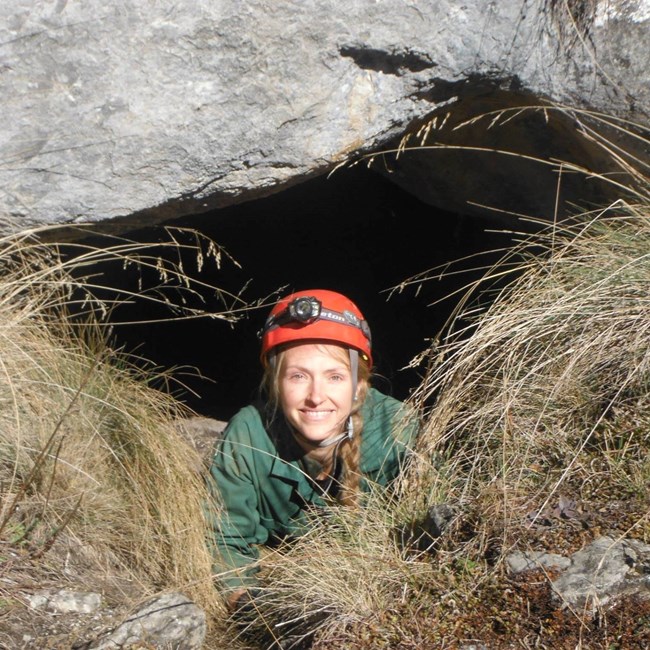 woman wearing a hard hat and headlamp sticks her head out of a cave opening