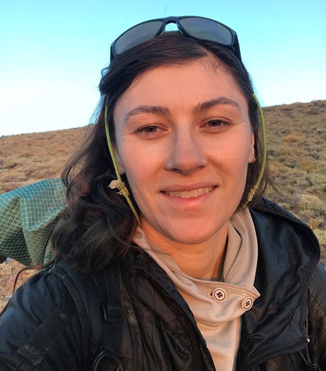 Closeup of woman outdoors with sunglasses perched on head and view of blue sky and shrubland behind her.