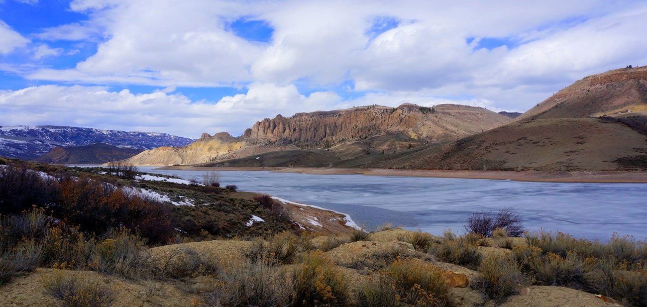 Blue Mesa Reservoir remains partly frozen in early spring below rugged cliffs in Curecanti National Recreation Area. Partially frozen reservoir below cliffs and rolling hills at Curecanti under a cloudy spring sky.