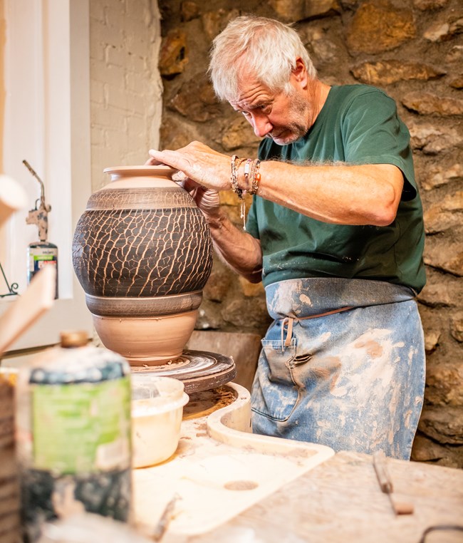 Potter wearing apron stands at table working on large ridged ceramic vessel with crackled texture.