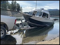 A silver pickup truck is backing a trailer with a boat down the paved boat launch ramp.
