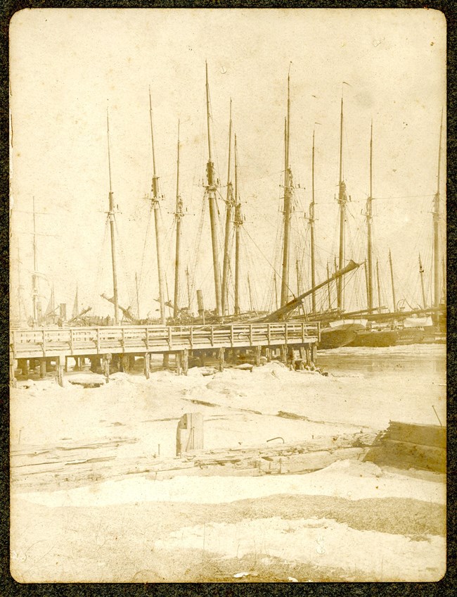 Photograph of mutiple sailboats docked at ice piers with a wooden dock in front.