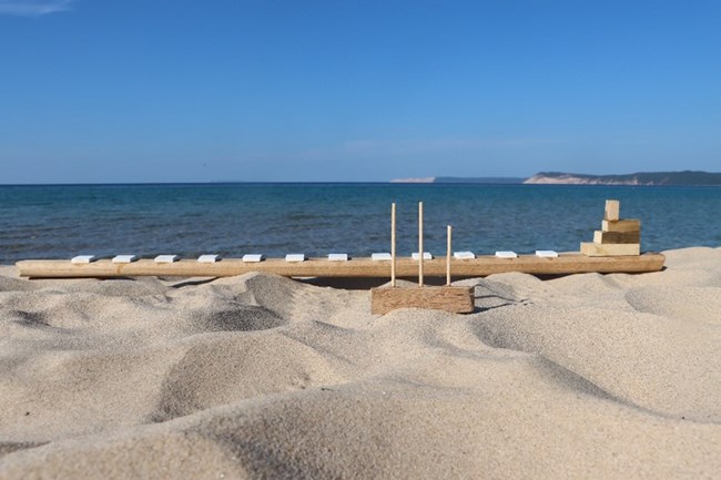 A sandy beach and purple-blue water. A large, pale brown toy boat with white hatches made out of wood takes up the majority of the frame lengthwise. In front of the boat is a much smaller boat, also made out of wood.