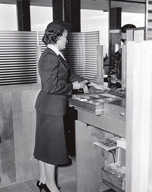 Bobbie Gleason A woman faces away from us towards a counter. She is wearing a dark colored uniform and pumps.