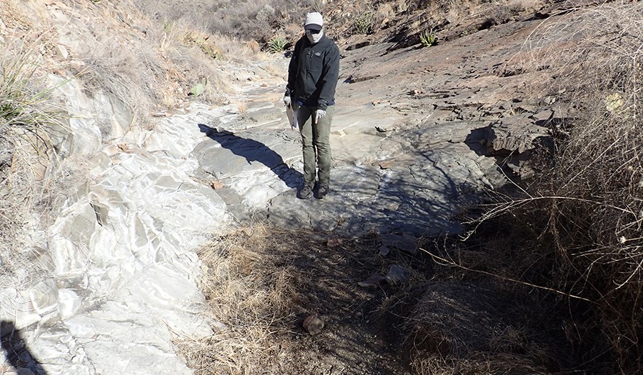 A person pointing at a dry depression filled with soil and dried plants next to bedrock in a drainage.