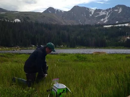 Man kneeling in a meadow in front of a lake and mountains