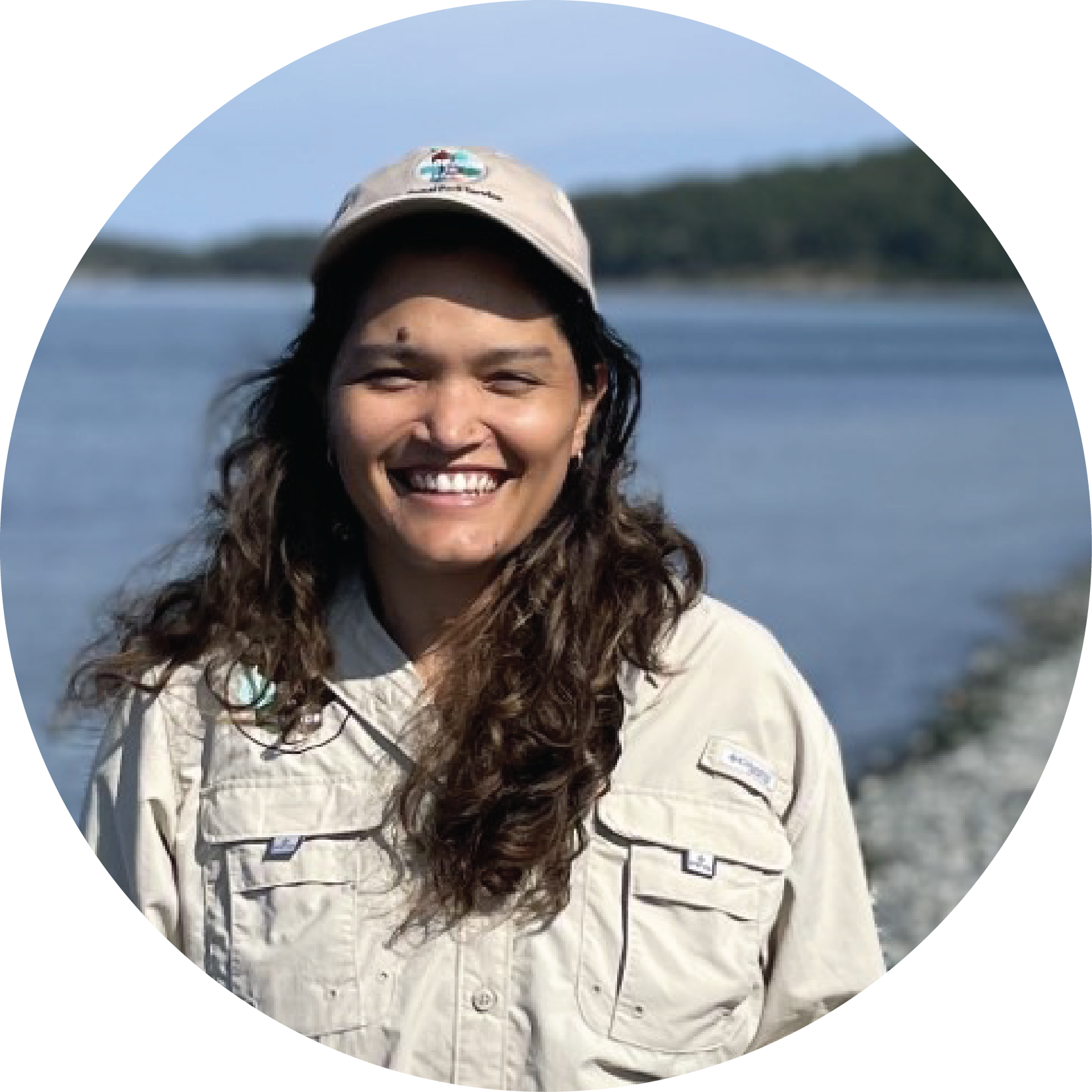 A smiling woman on a rocky coast with a khaki field shirt and ball cap.