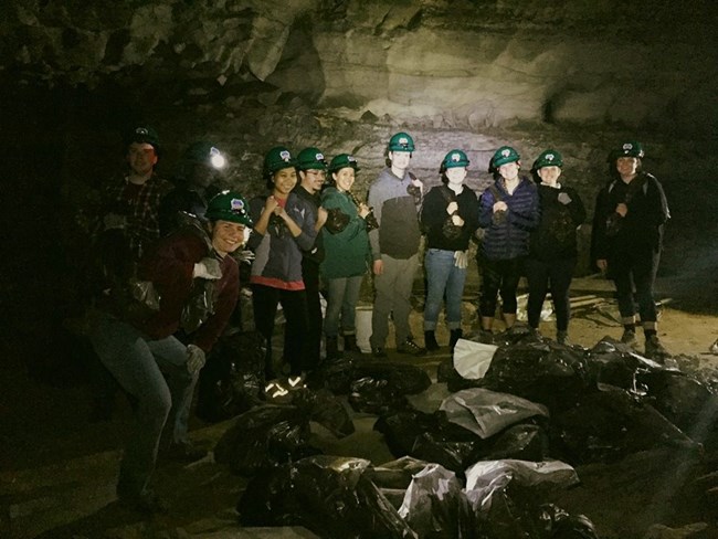 A group of eleven people wearing helmets and headlamps pose behind a pile of trashbags inside a dark cave.