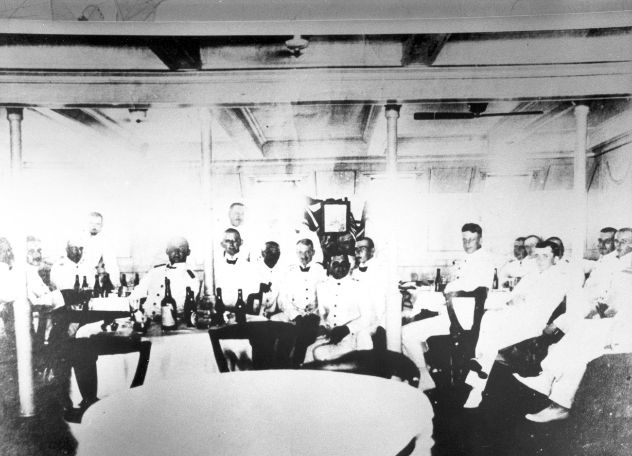Black and white photo of men in uniform sitting around a few tables