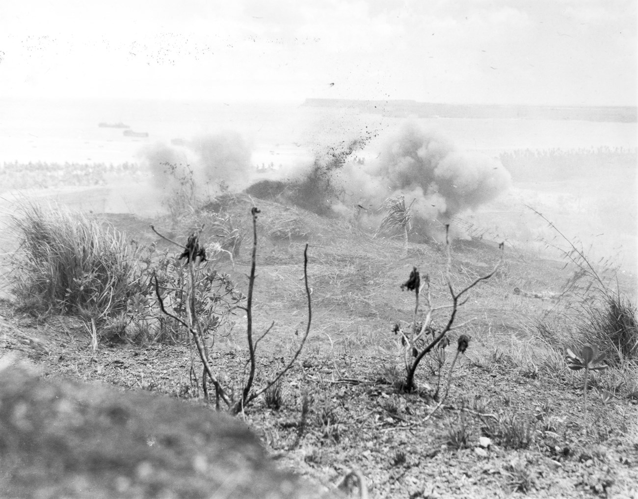 Imperial Japanese troops took advantage of established defensive positions in hillside caves as the Marines attacked the slopes of Mount Alifan. Marine engineers and demolition teams eliminated the cave positions with flamethrowers and grenades. A barren, battle-scarred hill with an explosion creating a cloud of smoke. Sparse vegetation in the foreground.