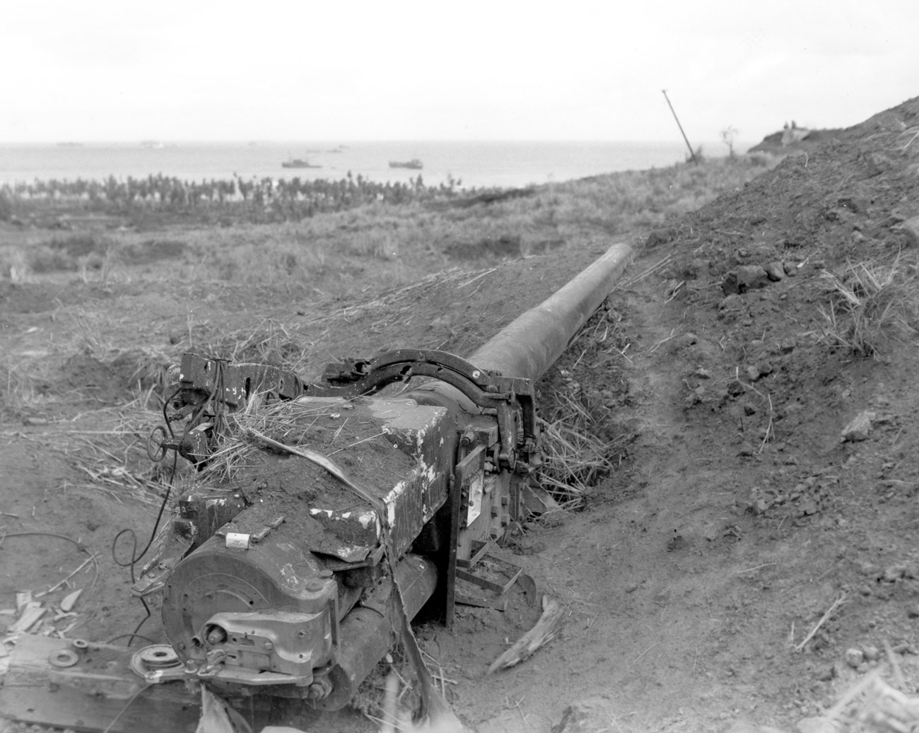 One of the Japanese 15.2cm coastal defense guns on Mount Alifan. The gun was located just 200 yards below the mountain's top and was trained on the landing beach. A rusted artillery piece lies in a muddy trench, overlooking a tranquil sea with distant boats and a crowd of soldiers in the background.