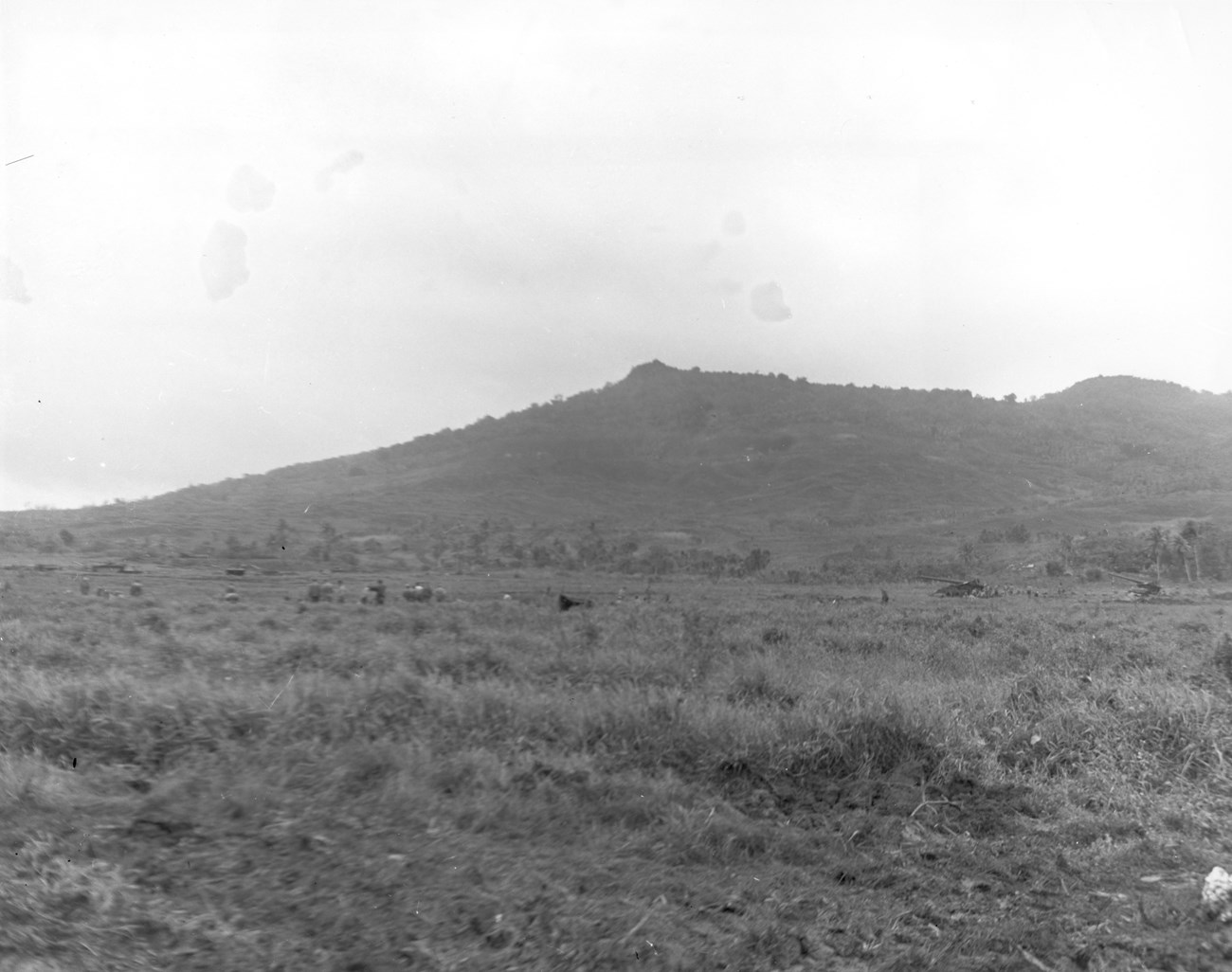 Mount Alifan, overlooking the Hågat Beachhead, was captured by U.S. Marines on July 22, 1944. In the foreground at the base of the mountain, Long Toms (155mm) field guns have been set up. A black-and-white landscape photograph shows an expansive grassy plain leading to a large, tree-covered hill under a cloudy sky. Large guns have been set up on the plain.