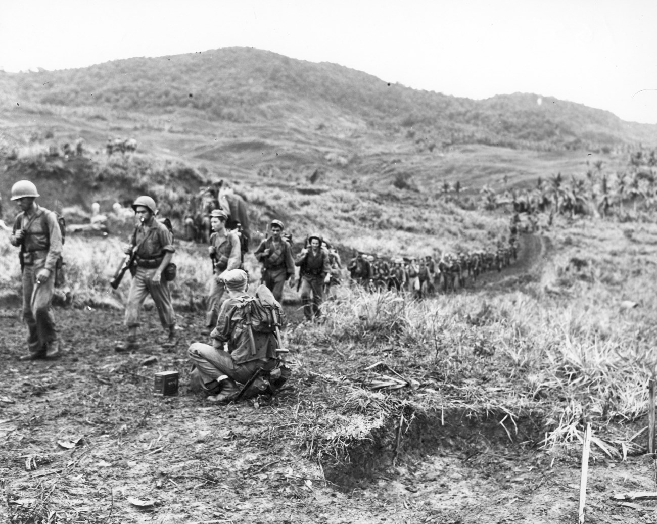 Members of the 4th Marines march out of the hills overlooking the Hågat Beachhead after successfully capturing Mount Taene (seen in the background). They had just been relieved by members of the Army's 306th Regiment, 77th Infantry Division. A long line of soldiers in helmets marches through a muddy, grassy landscape with rolling hills in the background.