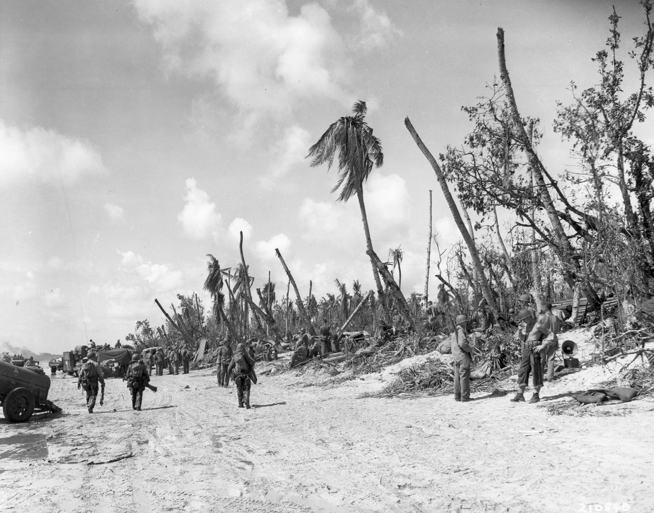The 305th Regiment, 77th Infantry Division finished landing on Hågat Beach the morning of July 22, 1944. Black-and-white image of soldiers on a war-torn beach, with damaged palm trees and military vehicles in the background.