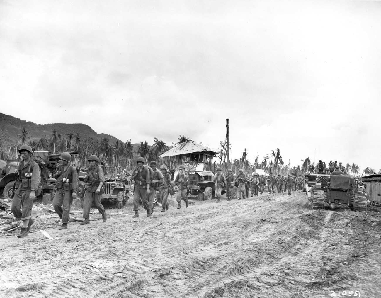 After landing on July 23, the 306th Regiments, 77th Division relieved the 4th Marines and assumed control of the entire perimeter east of Old Hågat Road. A group of soldiers in military uniforms march along a muddy road, with vehicles and a damaged building in the background amidst tropical trees.