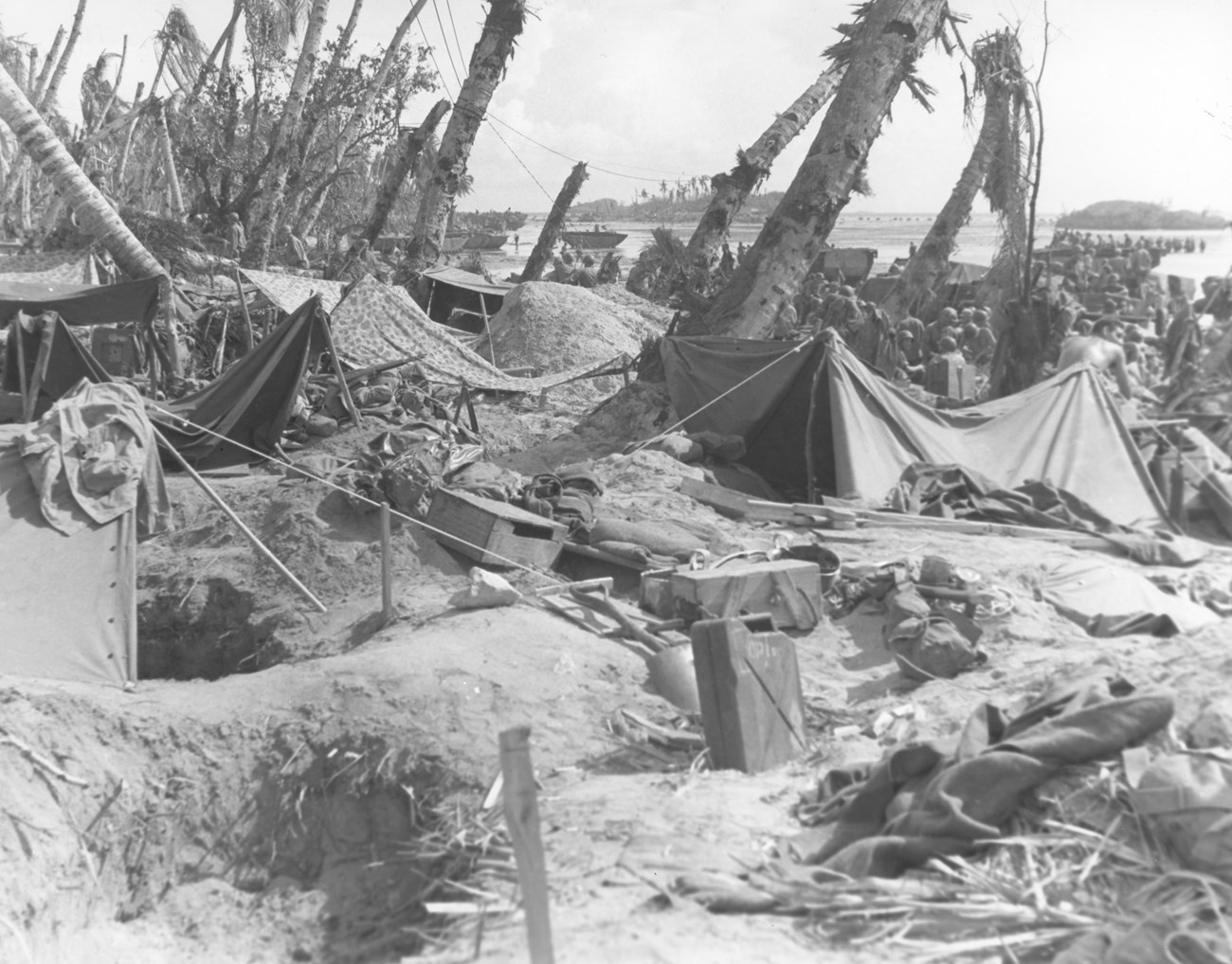 306th Regiment, 77th Infantry Division sets up a bivouac on White Beach at after wading to shore on July 23, 1944. World War II battlefield scene with tents, scattered supplies, and palm trees. Soldiers gather in the background.