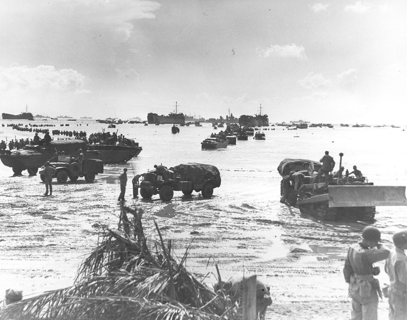 The 306th Regiment, 77th Army Division waded ashore the afternoon of July 23, 1944. Military vehicles and troops disembark at a beach, with boats and ships in the background under a cloudy sky.