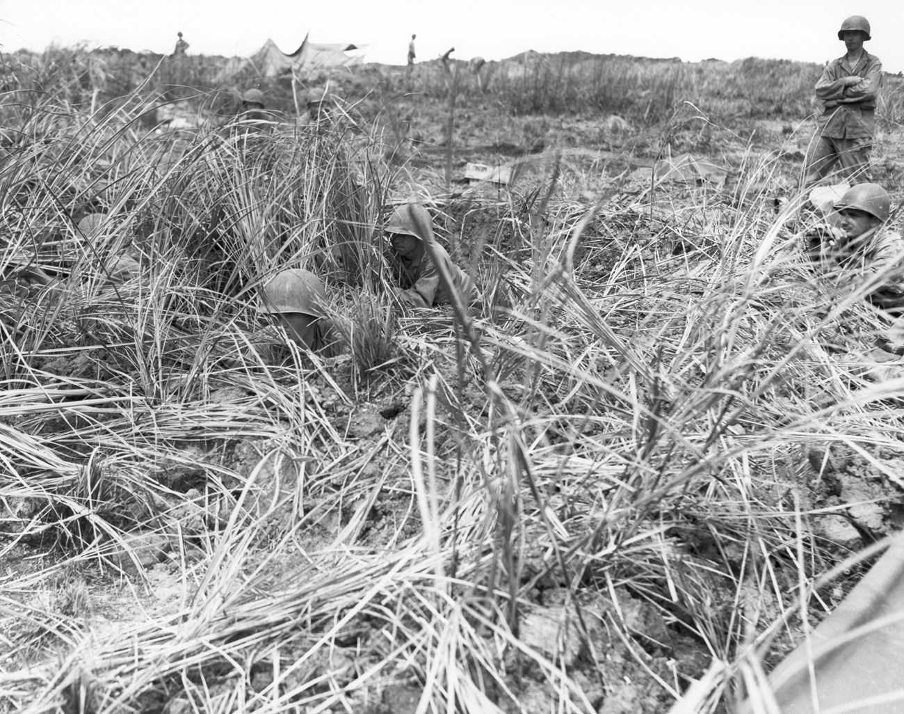 A U.S. machine gun nest on the left flank of the 306th Regiment, 77th Division, US Army during the second phase of combat in the Battle of Guam. Soldiers in helmets partially hidden by tall grass in a field engaged in a military operation. One stands with arms crossed.