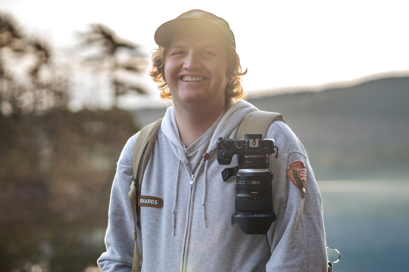 Brady Richards, Visual Information Specialist and Park Photographer, wearing a camera while hiking around Eagle Lake. Park ranger wearing a camera backpack in front of Eagle Lake.