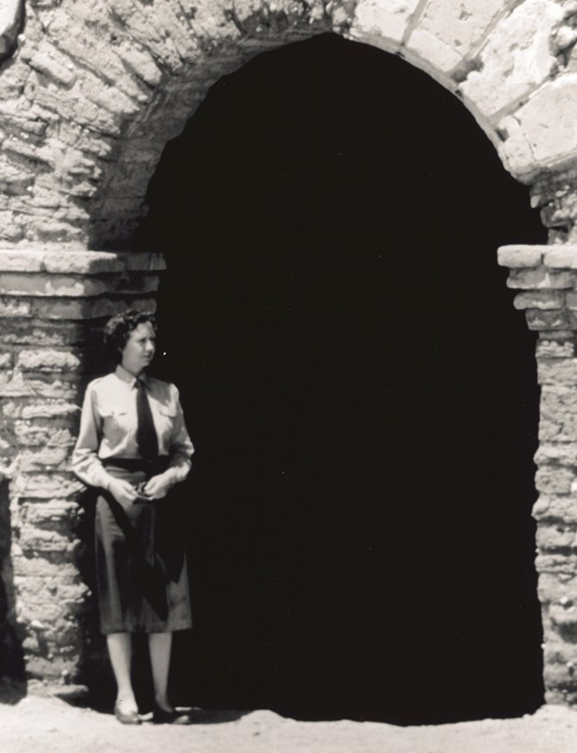 Sallie Brewer in NPS uniform with skirt stands in front of a stone building.