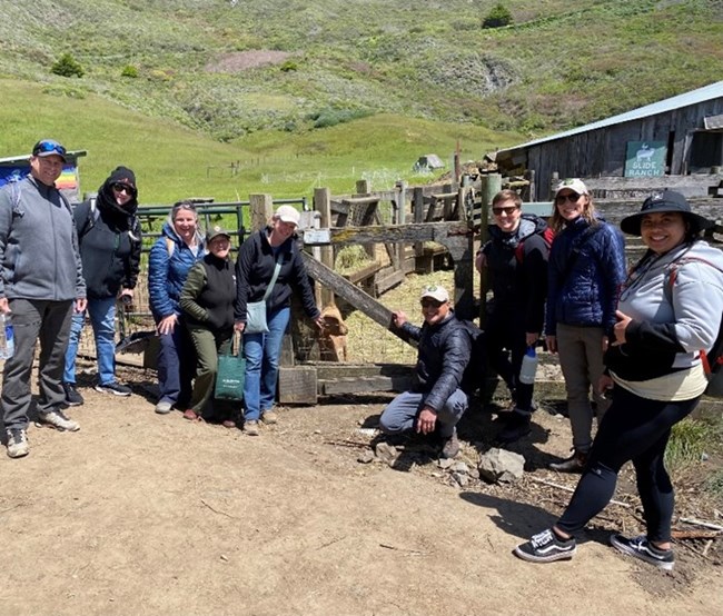 Part of the GGNRA Planning Division visiting a park site. Brian is kneeling next to the goat Group of individuals posing for a photo with a goat, a dirt path, and grass in the background.