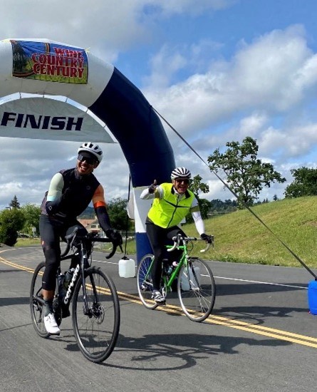 Brian Aviles, left, finishing a 100-mile ride in May Two males on bicycles. Male on left is wearing dark clothes, while male on right is wearing a bright green neon vest and dark pants. Behind them is a sign that says "finish," grass, and clouds.