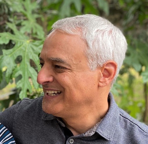 Profile Photo of Brian Aviles Older Latino male smiling with white hair looking off to the side with a grey shirt and trees in the background.
