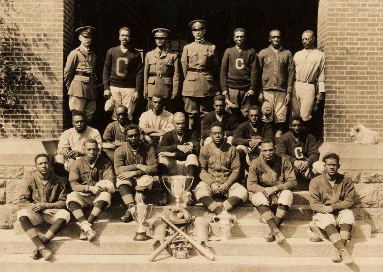 Image features 20 figures of which 18 are African American including: six in "C" sweaters; eight wearing a gray button up jacket w/"Cavalry" embroidered across chest; three wearing jerseys reading "Cavalry" and one of the three officers in top row. Photo