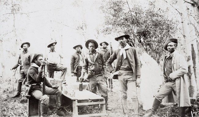 Eight African American men stand in a wooded area wearing worn clothes and wide-brimmed hats