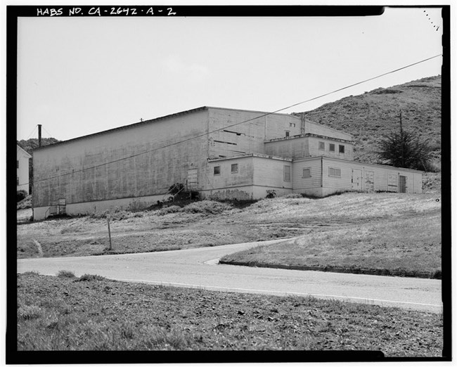 Perspective view of large, rectangular, multi-story building. The northern side has no windows and is covered in weathered light colored panels. The western side consists of three shorter attached tiered structures which each have a few windows.