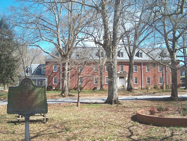 Metal sign on a vertical post reads Georgia: 1776: Georgia School For The Deaf. 14 lines of engraved text are illegible. The sign stands on a leaf-covered, grass field with scattered trees in front of a 2-story, red-brick building with 2 rows of windows