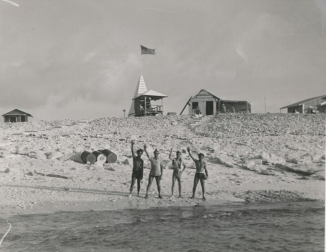 Black and white photo. Four men of color wearing shorts and hats stand at the shoreline. Behind them up a slope are four wooden structures. One flies the American flag.