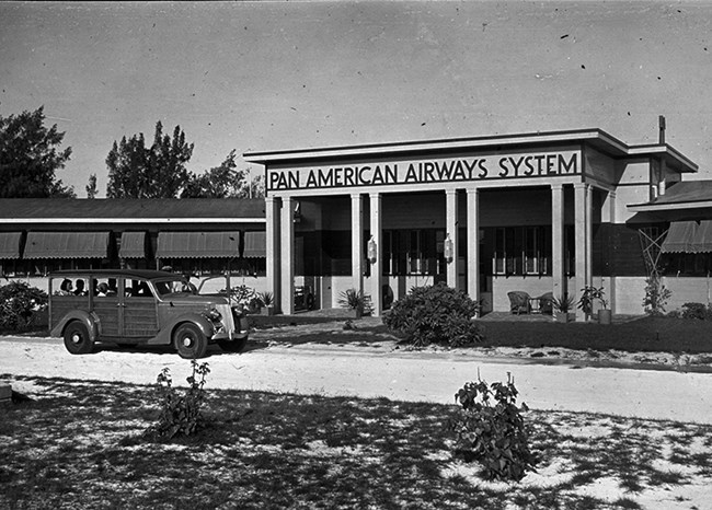 Black and white photo of a one-story building. The center entrance has “Pan American Airways System” over it. Several people sit inside a vehicle driving on a shell-covered roadway. There are small plants and scrub grass.