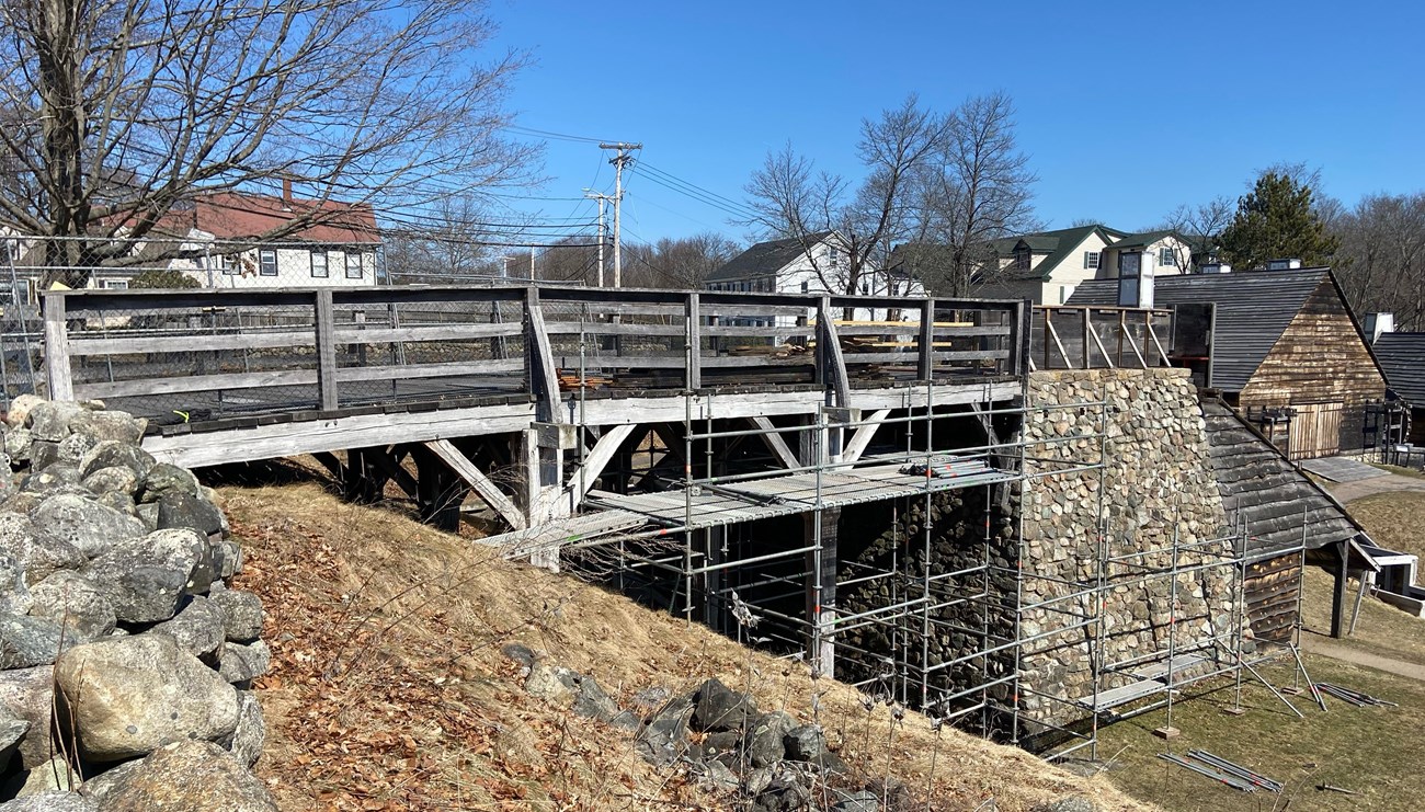 Wooden bridge leading to a large, stone furnace is surrounded by metal scaffolding on a cloudless day.