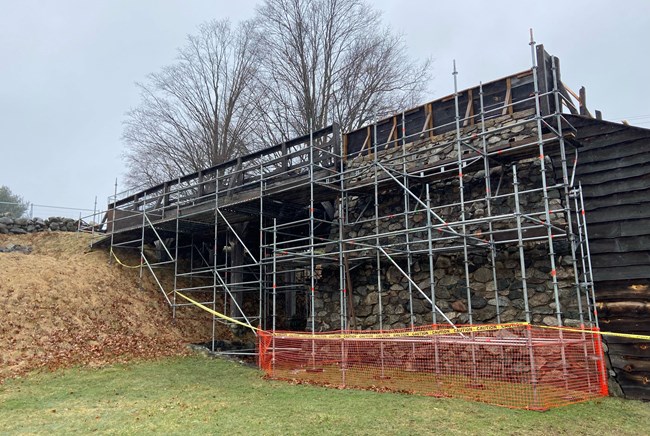 Metal scaffolding, orange fencing, and caution tape set up around a wooden bridge and stone furnace.