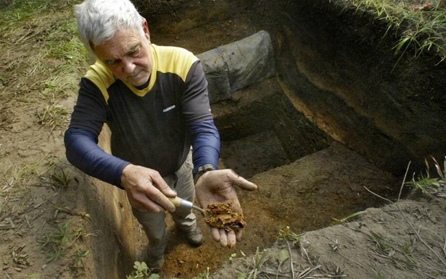 An older man with white hair sits in a rectangular archology dig. He is holding a brown artifact up to the camera.