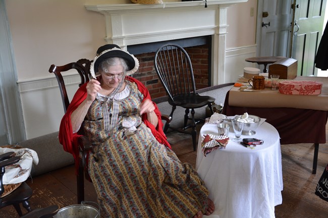 A woman dressed in 18th century clothing sewing in a chair.