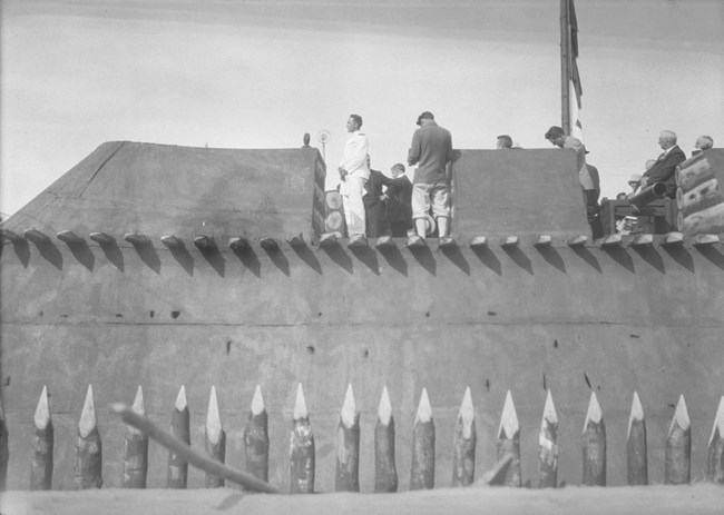 A man in a crisp white uniform stands in a miniature wooden fort addressing a crowd.