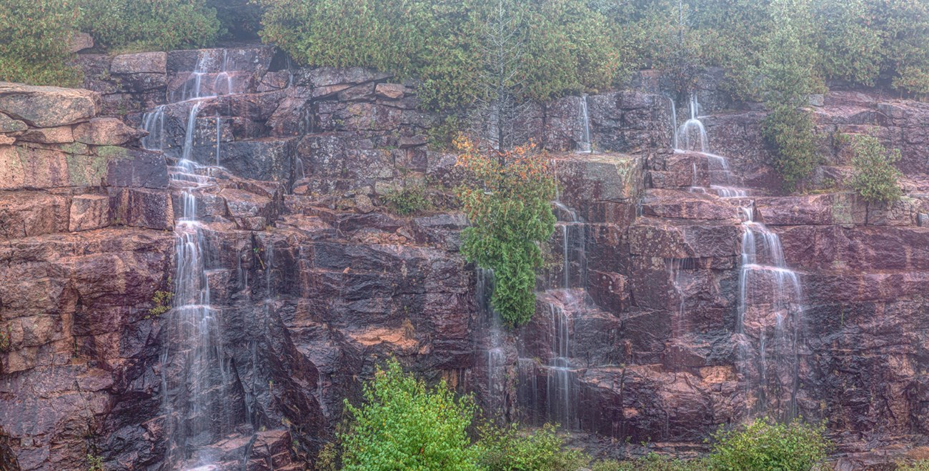Three separate small waterfalls trickle off a wide fractured cliff punctuated by green shrubbery Three separate small waterfalls trickle off a wide fractured cliff punctuated by green shrubbery