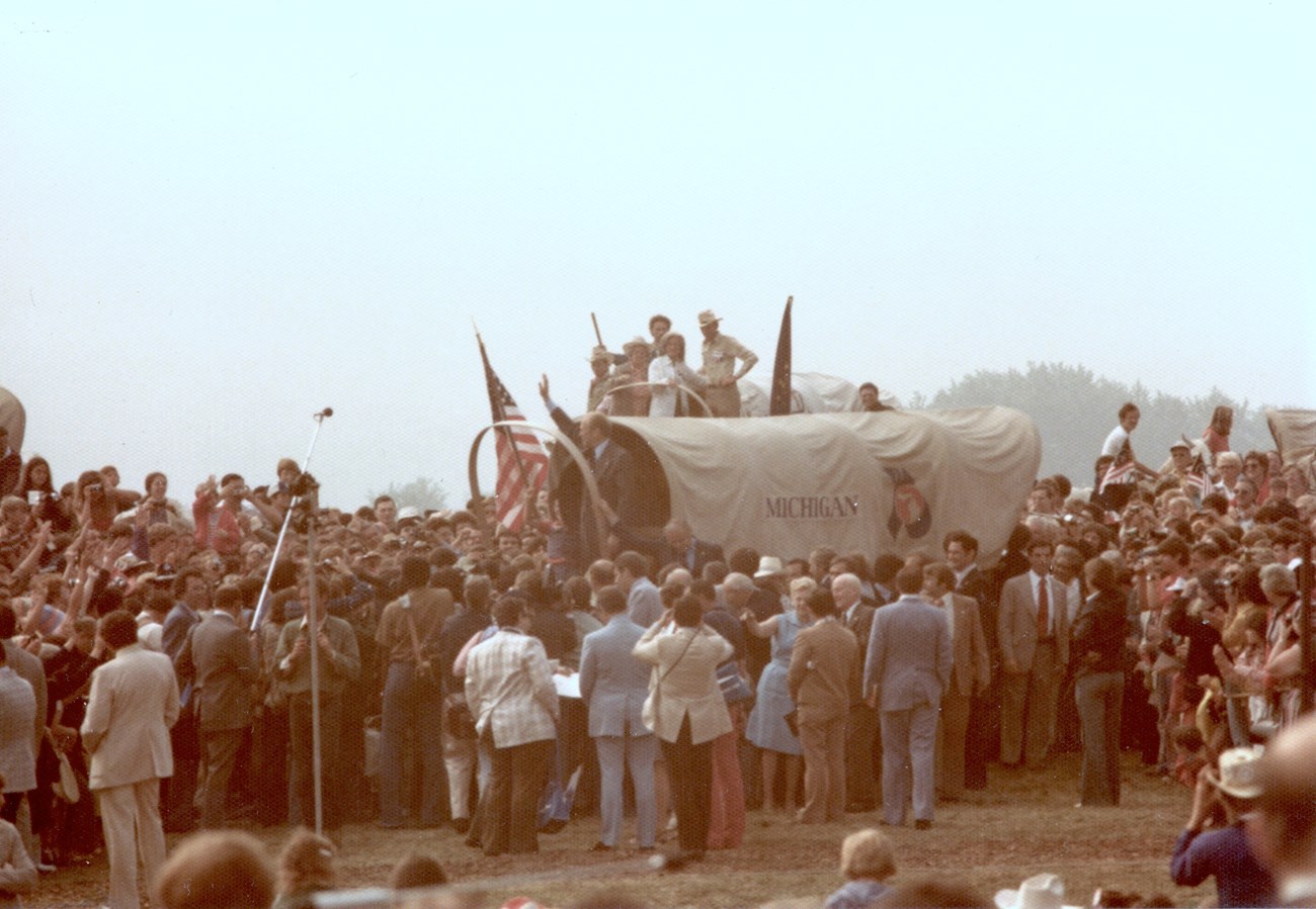 A person waves from a covered wagon surrounded by people.