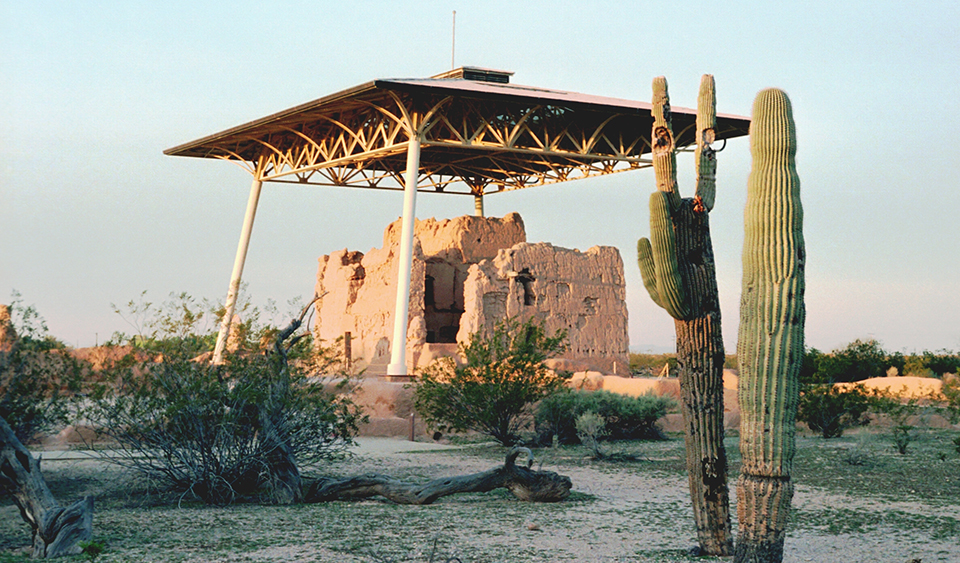 Partially eroded ruins of a massive adobe structure with protective modern roof structure overhead. The sun sets out of frame, bathing the structure in orange light. Two saguaros stand tall in front of the ruins.