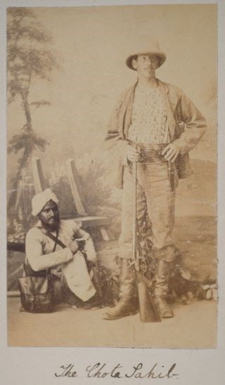 lt= Two men pose in front of painted scene, White man standing in boots holding gun, Sikh man wearing turban sitting