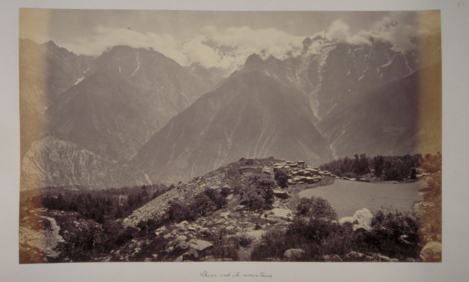 lt= Black and white photograph of small city in foothills of tall mountains with tops in snow and clouds