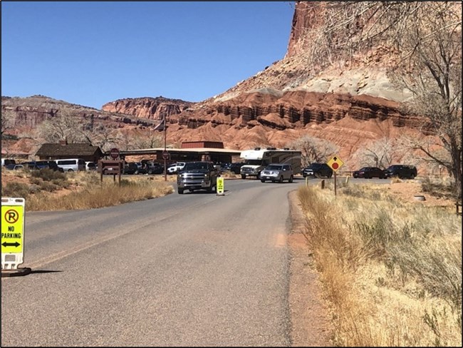 Capitol Reef Scene including cars on the road and in the parking lot at a visitor center, with colorful rock formations in the background