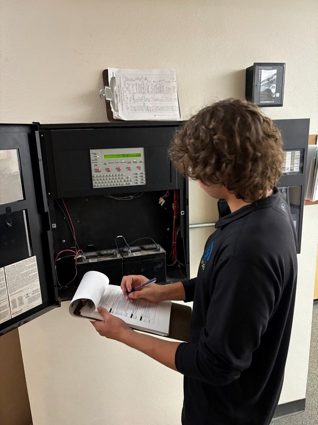 A  man holds a clipboard and looks at a readout from instrumentation on a wall.