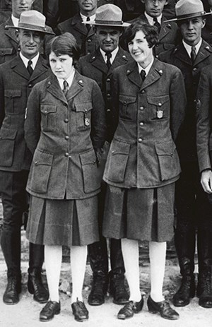 Two women in buttoned down jackets with wide pockets, pleated skirts, hosiery and oxford style shoes.