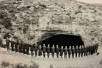 A line of uniformed rangers stand together on the outside of a cave opening in the rocks.