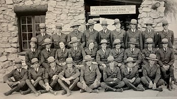 Three rows of rangers wearing uniforms pose in front of the Carlsbad Caverns museum.  THe front row is seated on the ground, the middle row sits on chairs and the third row stands.